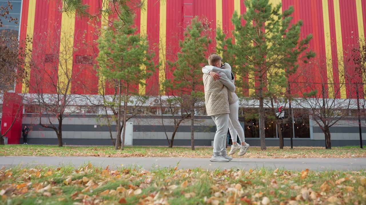 Joyful couple in matching outfits embrace warmly as man lifts and spins woman playfully amidst autumn scenery with colorful striped building and swaying trees, people passing by