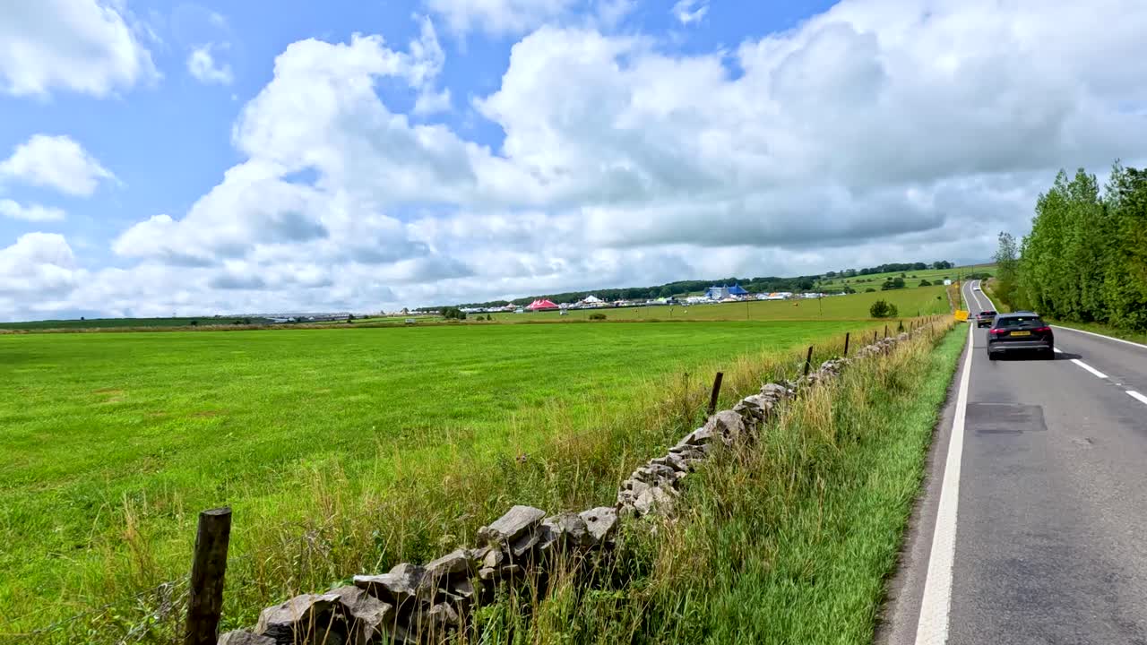 A car travels down a winding rural road bordered by stone walls and green fields under bright daylight with scattered clouds, captured from a moving vehicle