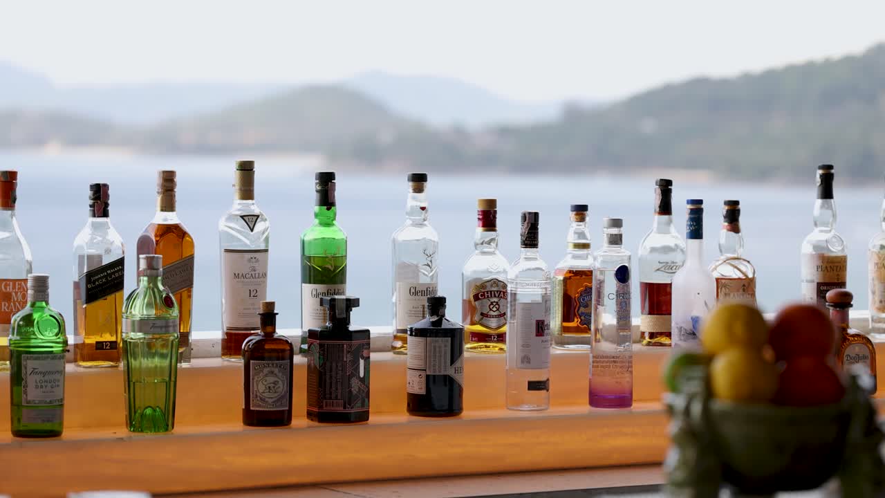 una vibrante variedad de botellas de licor en un mostrador de un bar de playa con un paisajístico telón de fondo de phuket. la luz del día brillante mejora la escena colorida