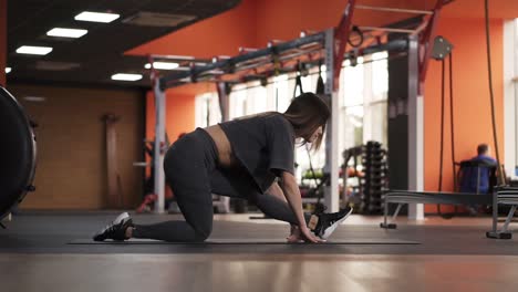 flexible skinny girl stretches before the split, doing the split, stretching to left leg and makes a slope forward on the mat in the modern, stylish gym. side view