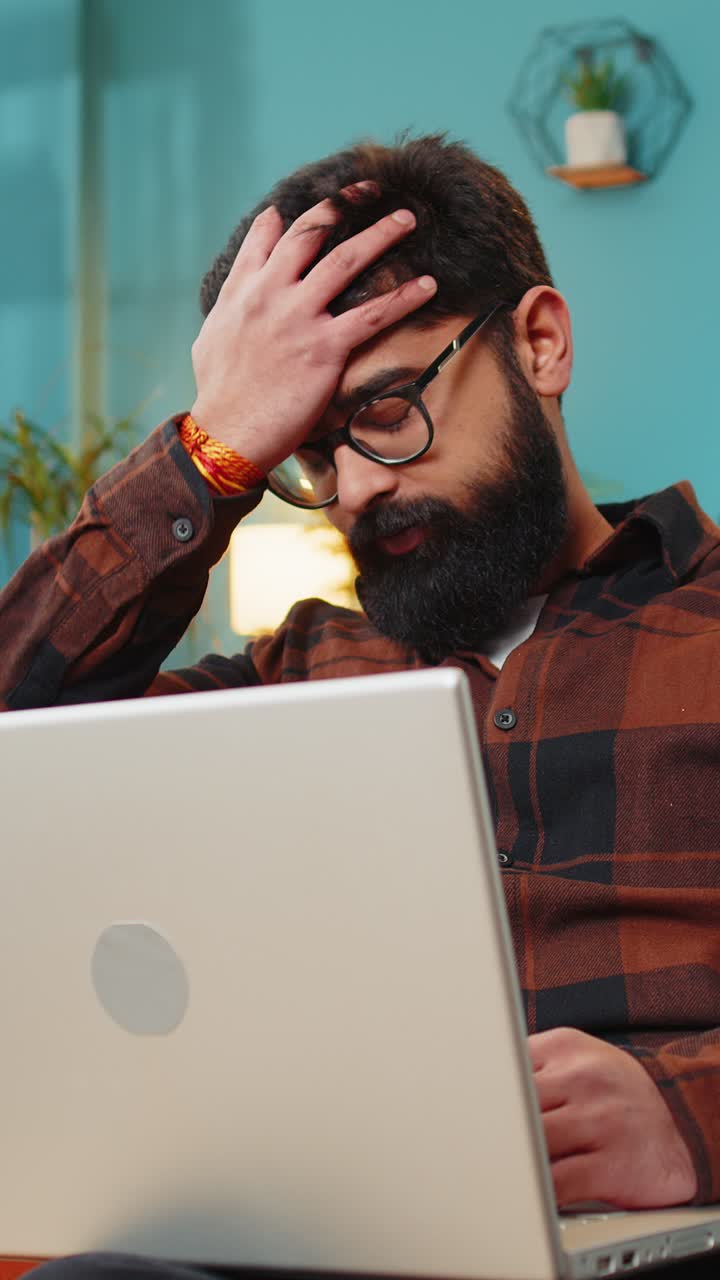 Upset indian man sitting on home sofa using laptop having anxiety stress loss bad fail news virus