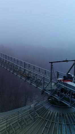 Sloping roof of an elevator tank with massive metal support over it. Drone footage along the metal pillar on the cistern. Foggy winter forest backdrop. Vertical video