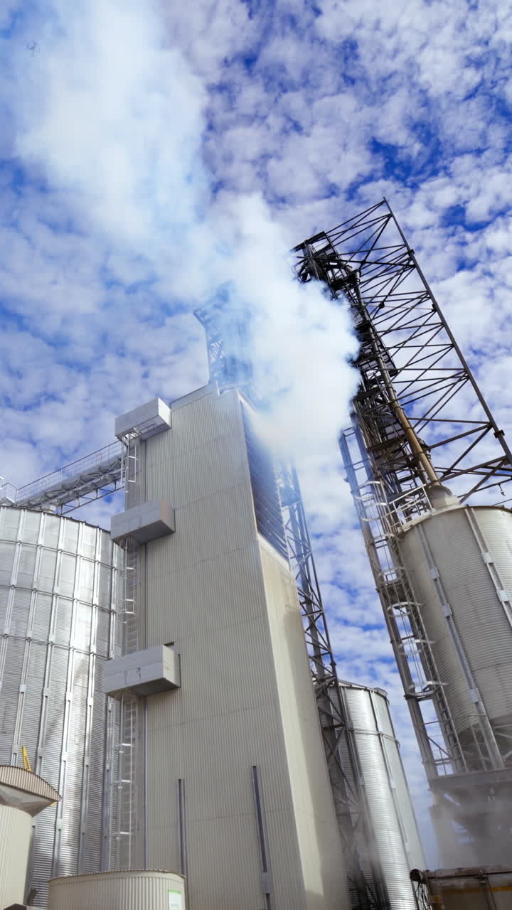 Modern factory for grain storage. Massive smoke from industrial plant. Large silver containers for drying grains. Pollution the environment. Vertical video