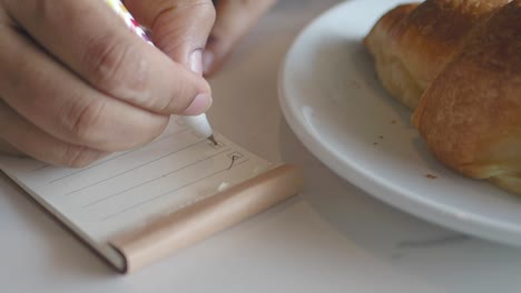 una mujer escribe en una lista de comprobación con un bolígrafo mientras disfruta de un croissant