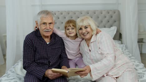 Cute-girl-with-senior-retired-grandmother-and-grandfather-sitting-on-bed-reading-book-in-bedroom