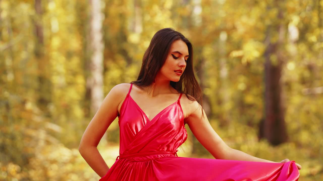 Una joven elegante con un vestido rojo vibrante posa con gracia en medio de un hermoso paisaje otoñal lleno de follaje dorado y una suave iluminación natural.