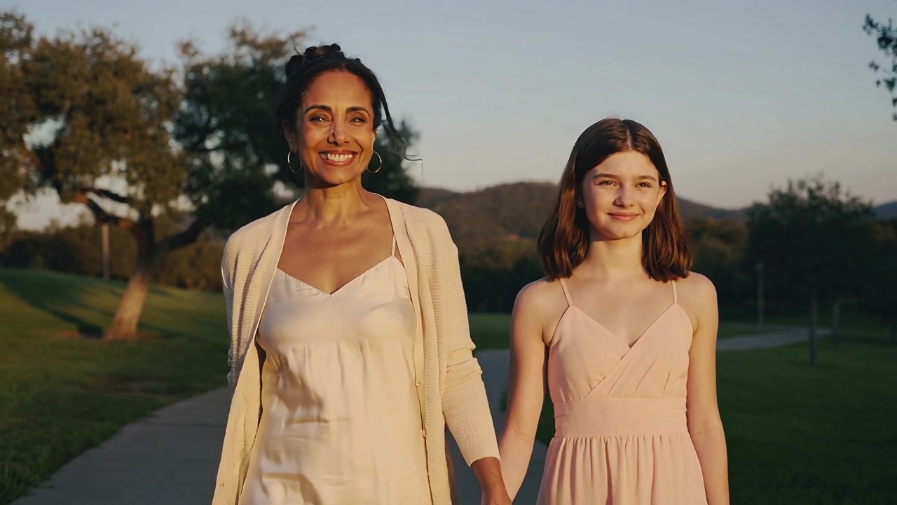 Mãe e filha caminhando de mãos dadas em um caminho no parque durante a hora dourada, vestindo cardigã e vestido rosa.