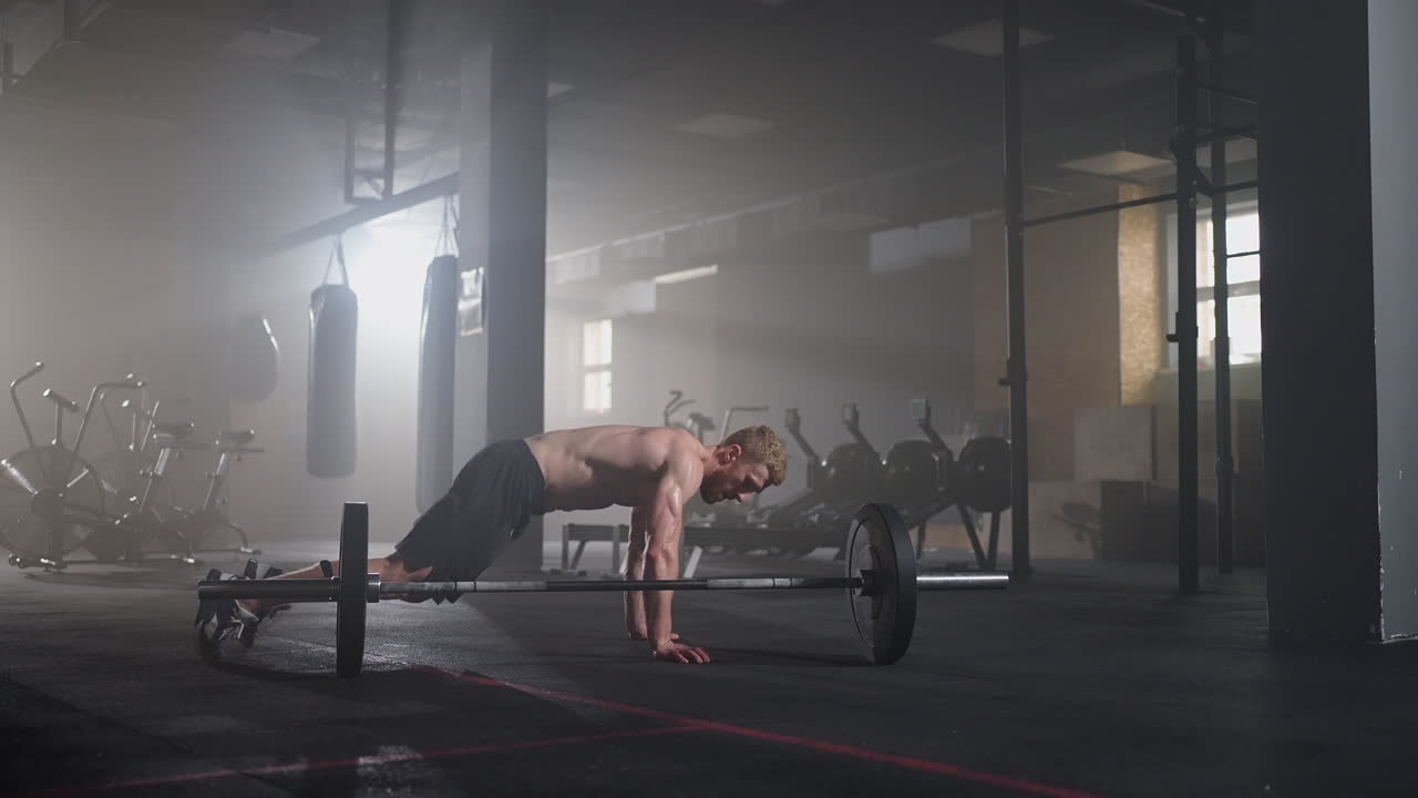 En ung man med bar överkropp i slowmotion gör en burpee över en skivstång på gymmet i motljus