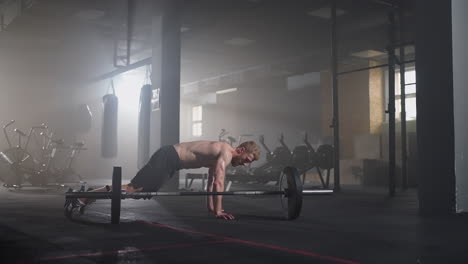 Un-Joven-Sin-Camisa-En-Cámara-Lenta-Salta-Un-Burpee-Sobre-Una-Barra-En-El-Gimnasio-A-Contraluz