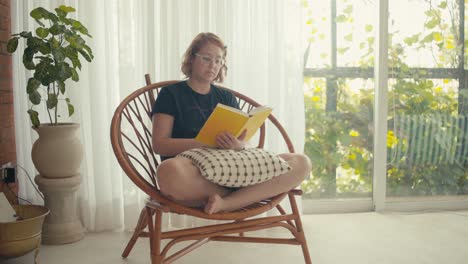 bright indoor scene with young woman on rattan chair reading a book, medium shot