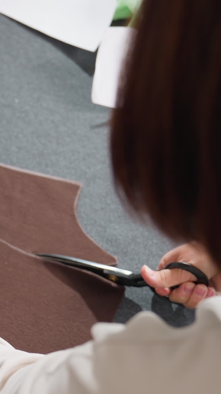 Caucasian woman cutting brown fabric on gray table using large shears, tracing chalk lines, focused hands shaping pattern, closeup of tailoring workshop and precise patternmaking process