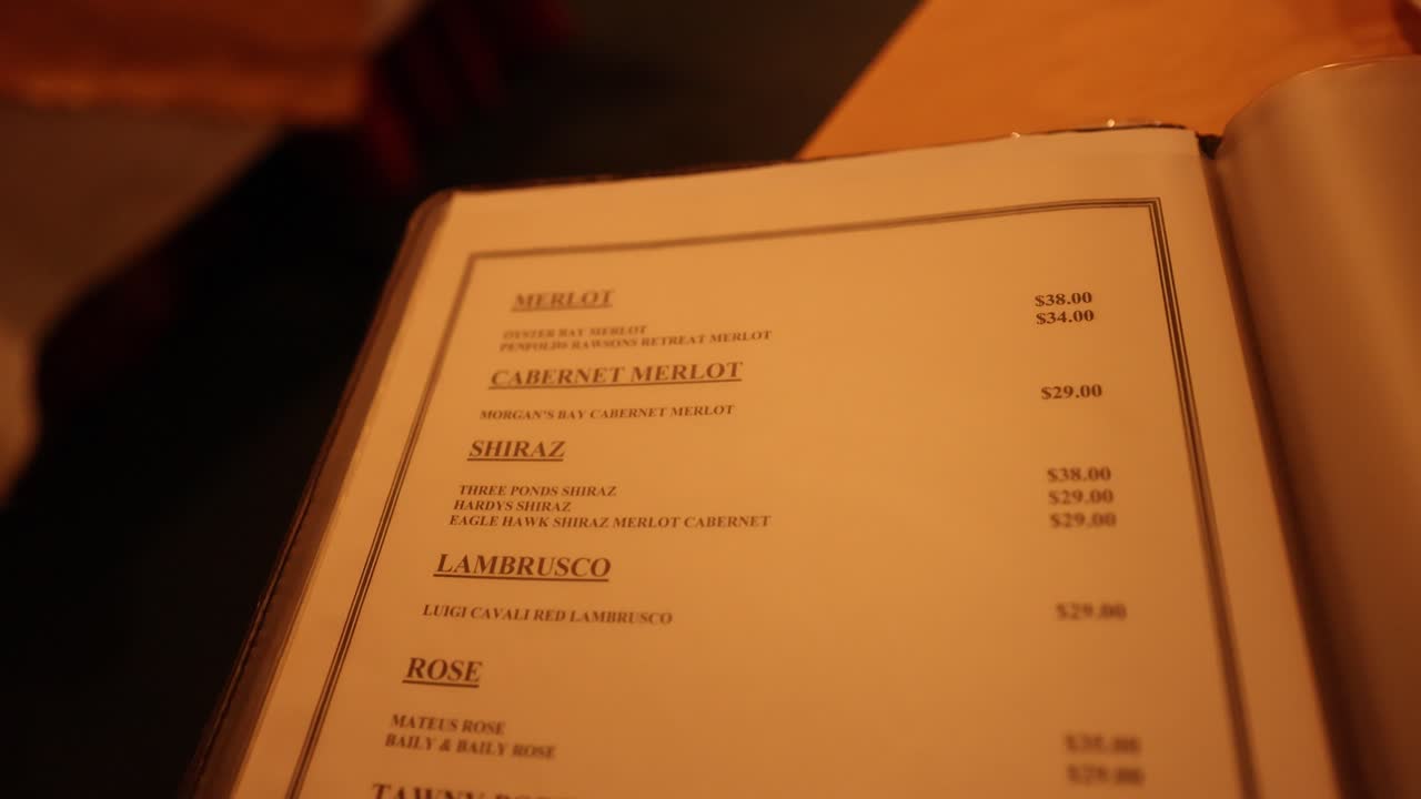 Person flips through wine menu under warm lighting in Australian restaurant, close-up handheld shot