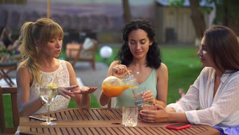 three female friends sit in the outdoors cafe, drink juice and have fun communicating, clinking with glasses