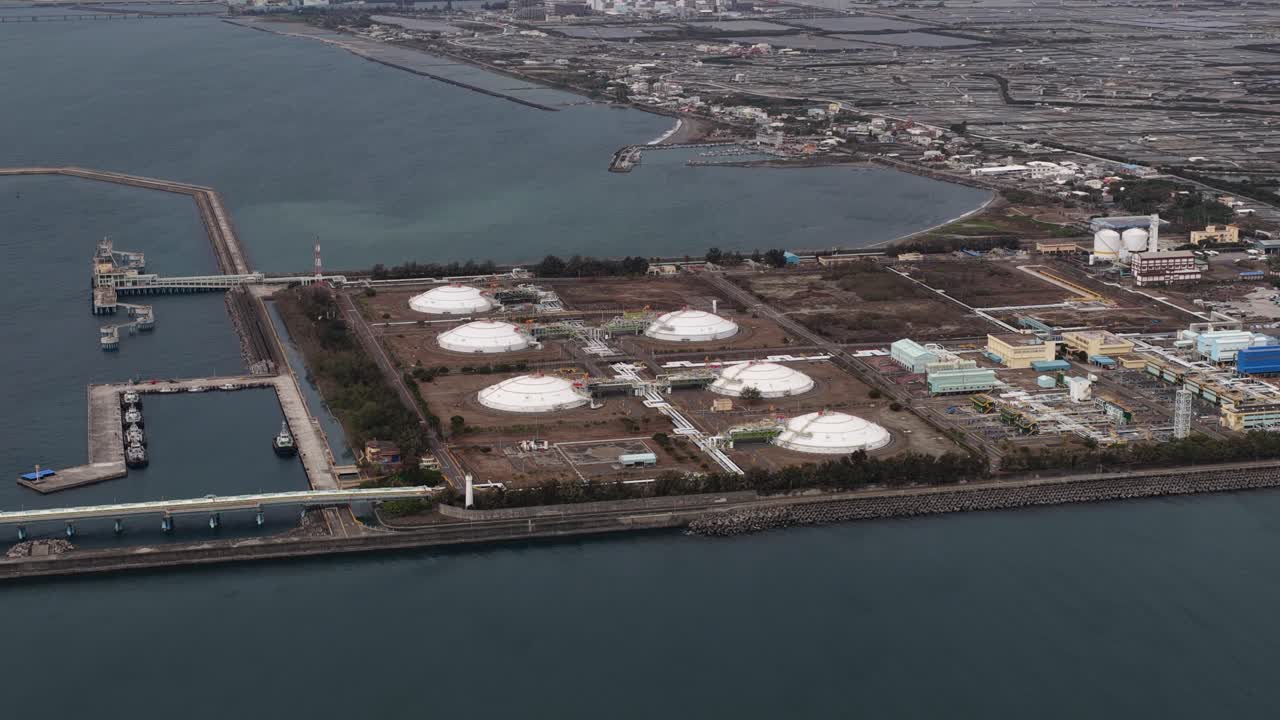 Veduta aerea da un aereo di un terminale di gas naturale liquefatto (GNL) in una zona industriale costiera, con grandi serbatoi di stoccaggio, un porto per navi cisterna e una città circostante in una giornata nuvolosa.