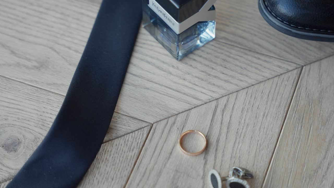 Close-up shots of groom's wedding accessories including tie, cufflinks, perfume, and a gold ring, neatly arranged on a wooden floor