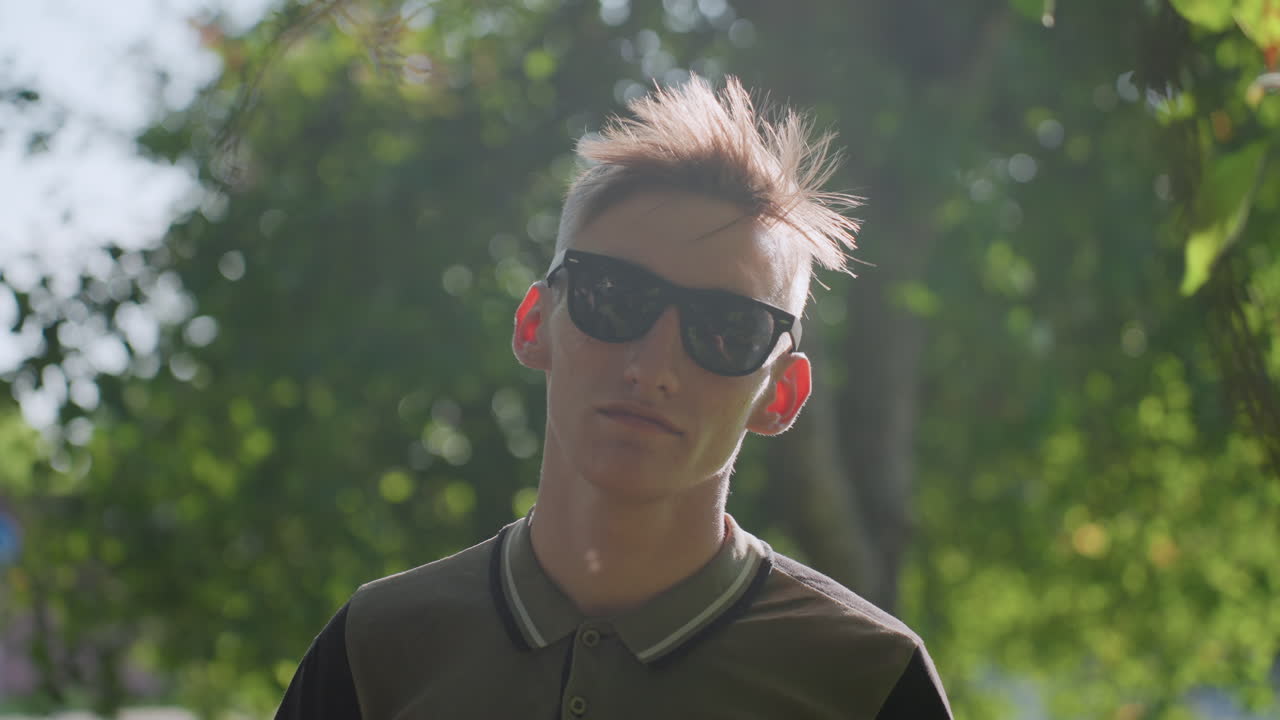 Teen Man In Sunglasses Tilting Head With Playful Defiant Attitude, Spiky Hair Backlit By Afternoon Sun, Leafy Park Background, Casual Polo And Confident Stare Conveying Rebellious Energy