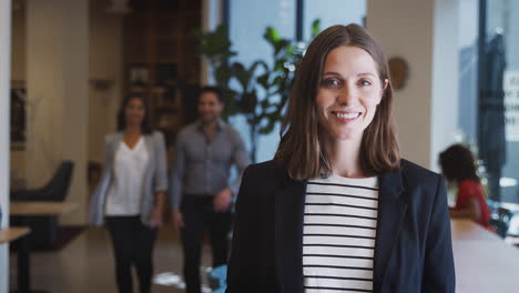 Portrait-Of-Businesswoman-Standing-In-Busy-Modern-Open-Plan-Office--With-Colleagues-In-Background