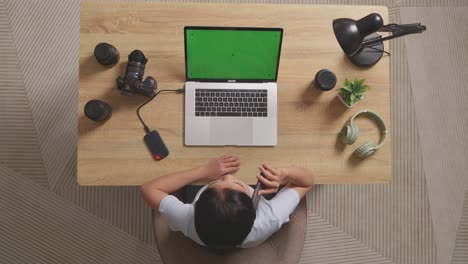 top view of a woman video editor talking on smartphone while using a green screen laptop next to the camera in the workspace at home