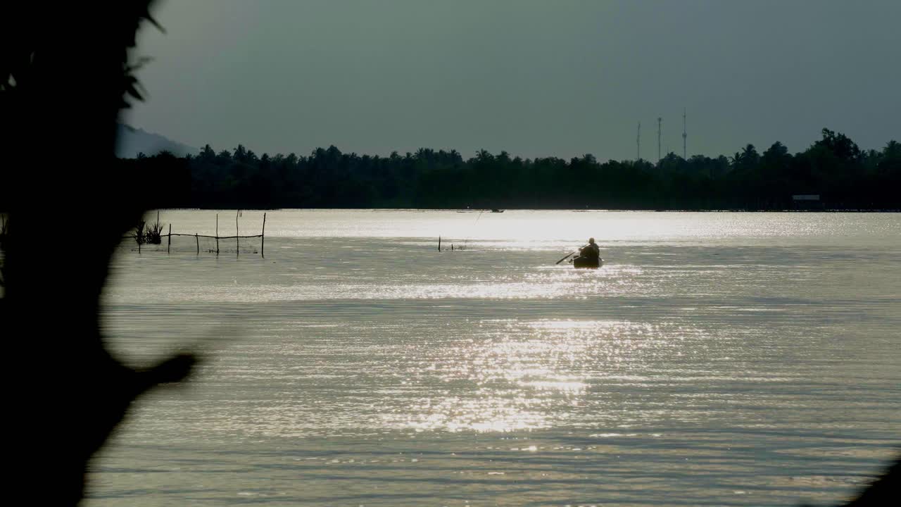 An abstract silhouette shot of a local Thai fisherman paddling a boat towards the bank of the Chanthaburi River, in the distance the beautiful morning sunrise reflecting off the water, Thailand