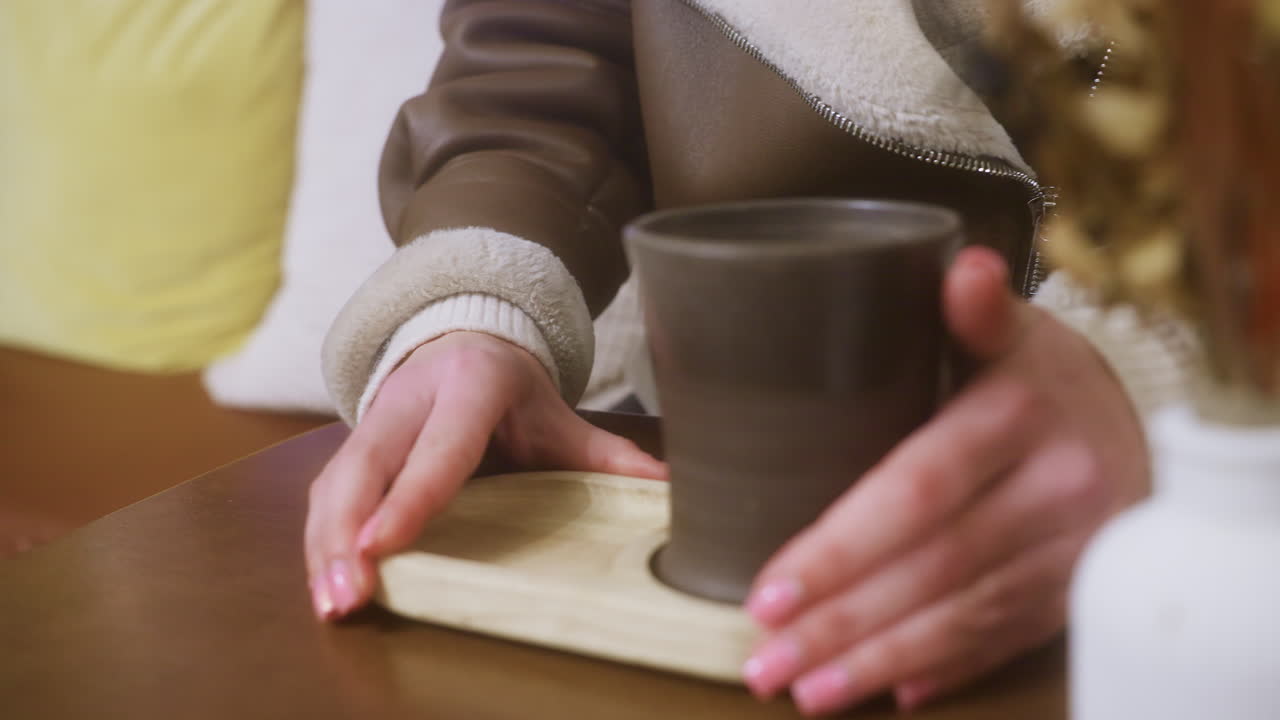 Close-up of female hand in brown shearling jacket holding cup of coffee in cozy cafe. Soft ambiance with blurred window view, creating a relaxed and inviting atmosphere