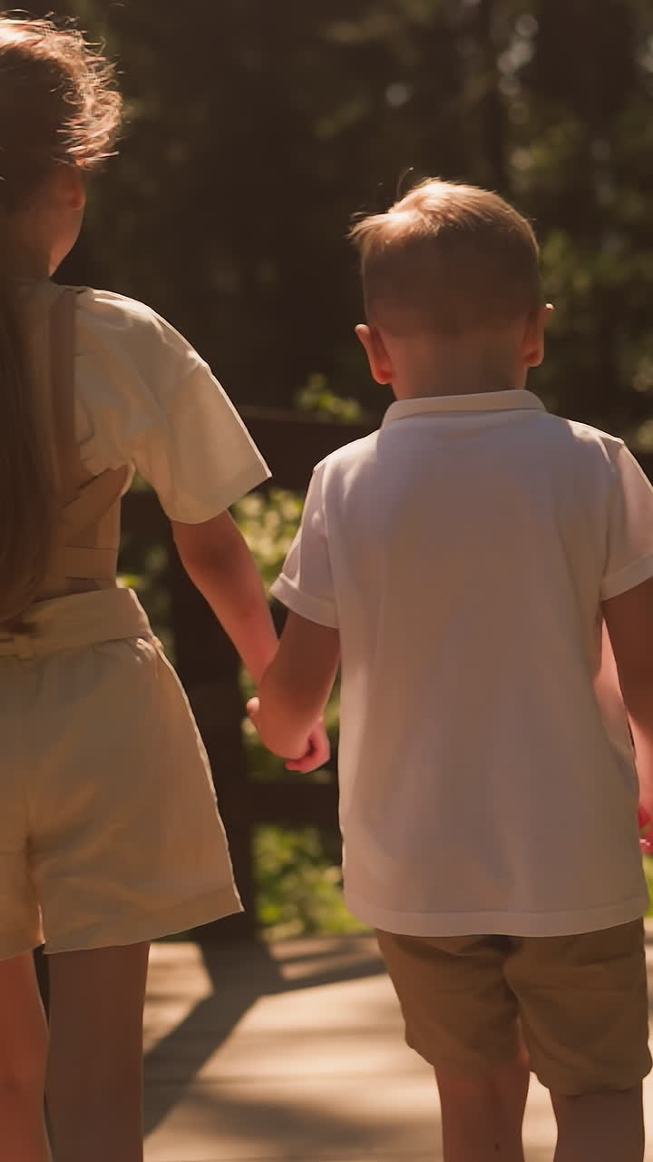 Little brother and sister walk joining hands along wooden footbridge in forest. Happy kids spend summer vacation at countryside. Children in park