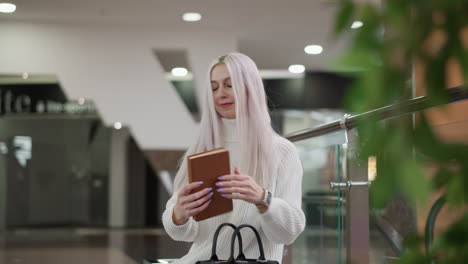 modern lady smiles walking to bench beside moving walkway, retrieves hardcover book from sleek bag, delicately flips pages and reads against blurred tree decoration backdrop in mall interior