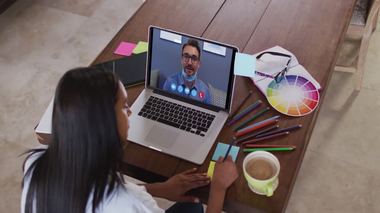 Mujer caucásica usando una computadora portátil en una videollamada con un colega y tomando notas.