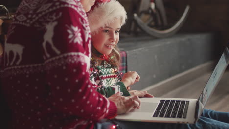 Niña-Feliz-En-Suéter-De-Navidad-Y-Sombrero-De-Santa-Sentada-En-El-Suelo-Con-Su-Madre-Y-Mirando-Algo-En-La-Computadora-Portátil