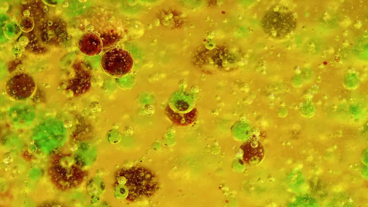 Close-up view of green and brown droplets interacting dynamically within a yellow liquid environment.