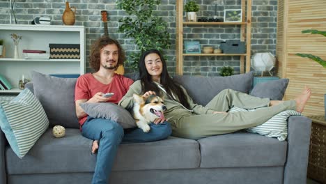 young family and corgi dog watching tv on couch at home laughing relaxing