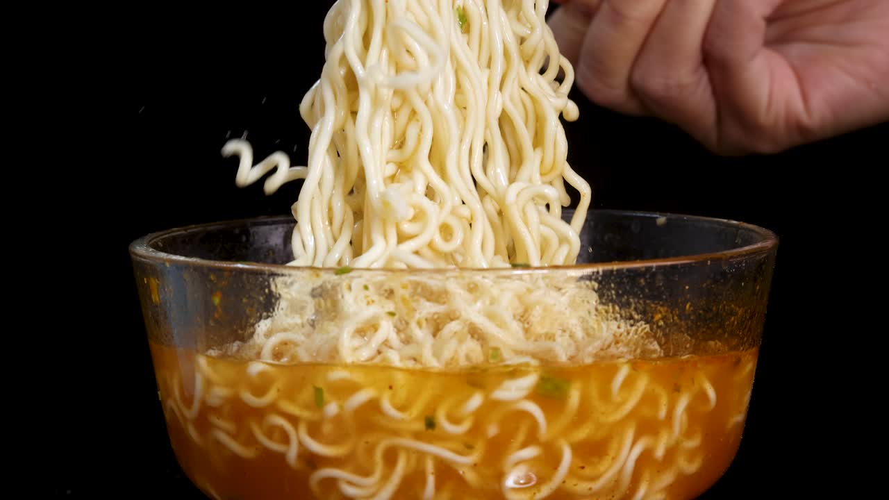 A fork lifts cooked instant noodles from a glass bowl of broth against a black background, with dramatic studio lighting and a static camera angle