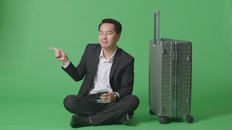 짐과 여권으로 가득 찬 아시아 사업가 여행자의 몸은 녹색 스크린 배경 스튜디오에 앉아있는 동안 웃고 을 가리키고 있습니다.