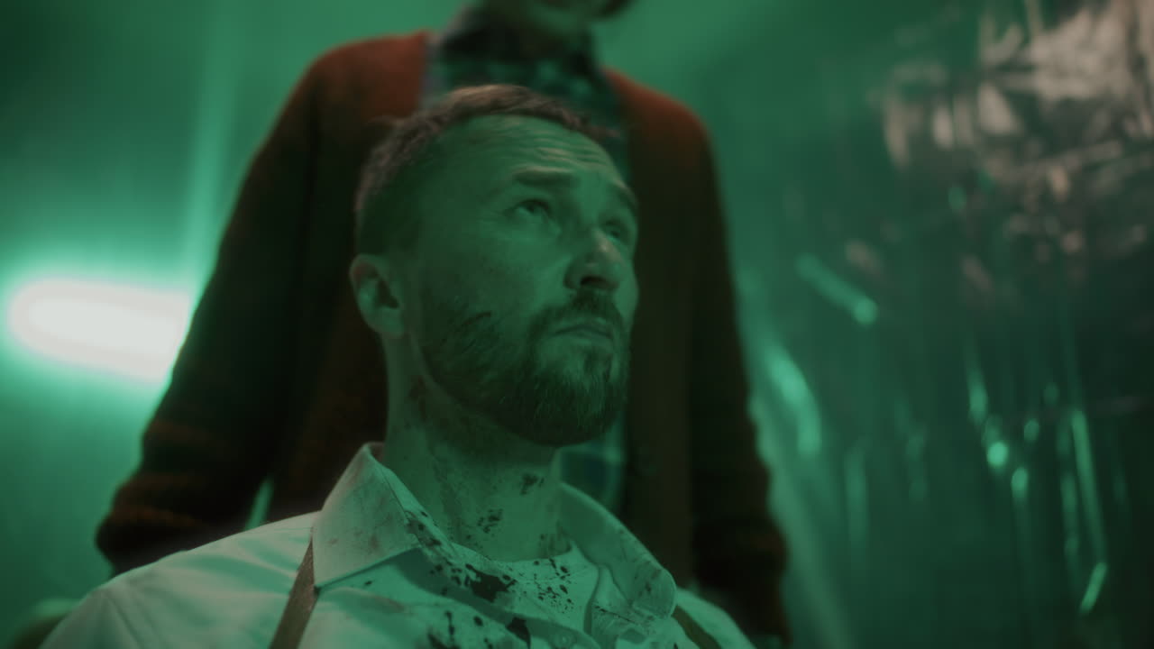 범죄자가 어두운 지하실에서 피를 흘린 인질의 머리에서 질 봉투를 제거하고 있습니다.