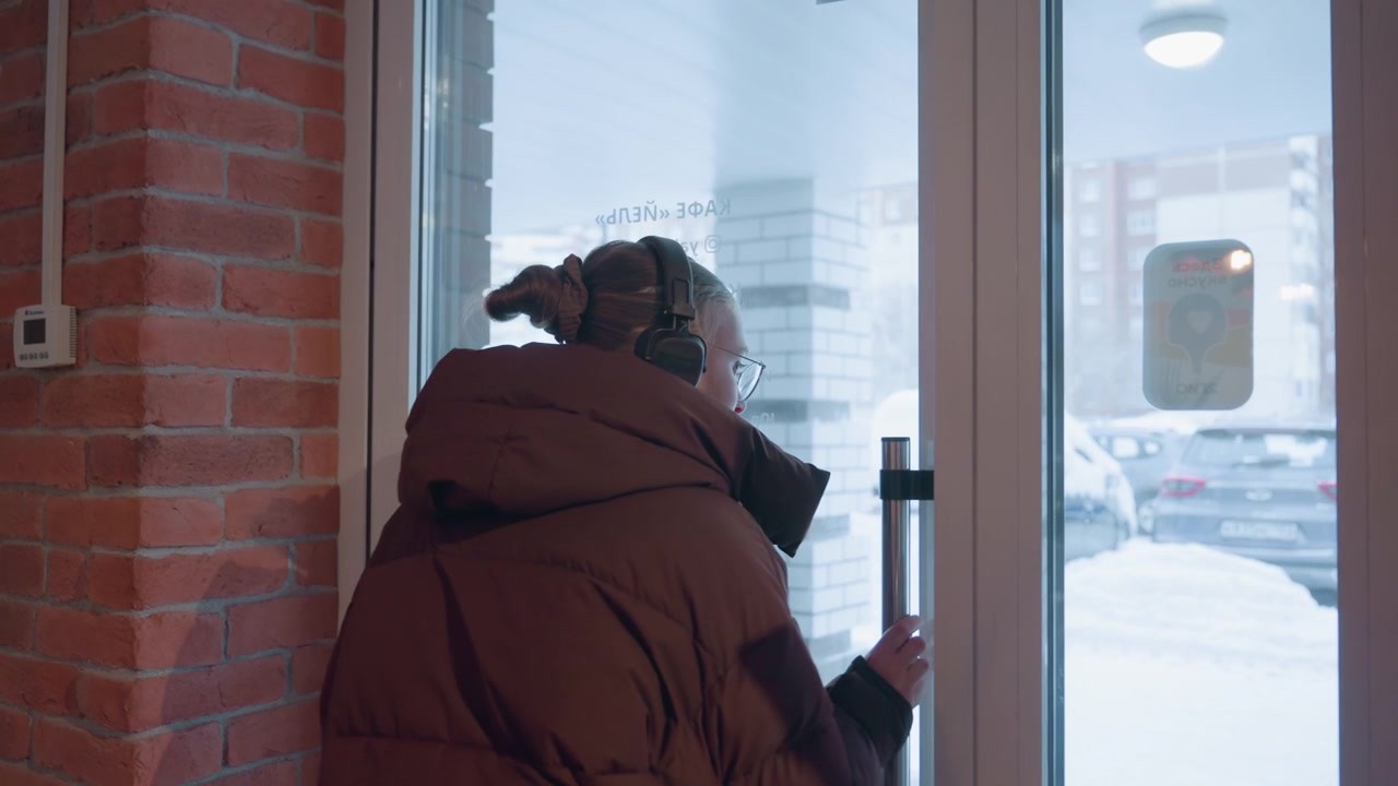 Woman in Winter Coat Exiting Building Through Glass Door to Snowy Street