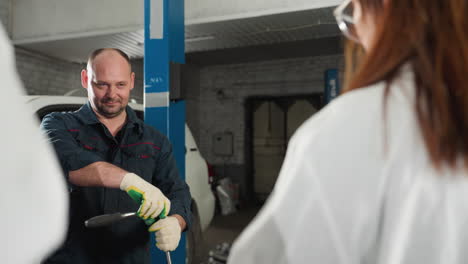 engineer demonstrates spanner use on car engine while students observe in mechanical workshop, mechanic smiles while handling tool, industrial workspace includes vehicles, and equipment in background