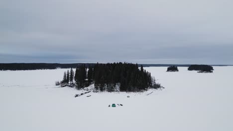 a drone out wide landscape shot of an island on frozen canadian paint lake with an ice fishing hut and skioos