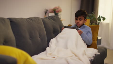 young boy sitting under blanket on sofa at home playing game or streaming onto handheld gaming device