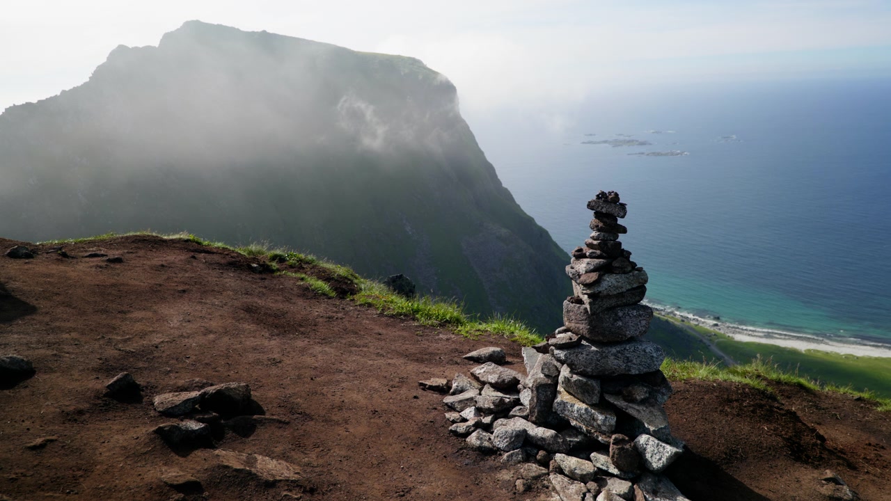 Ometto di pietra su un'alta vedetta sterrata sopra un ripido pendio costiero con spiaggia e mare aperto che sfuma nella nebbia nelle Lofoten.