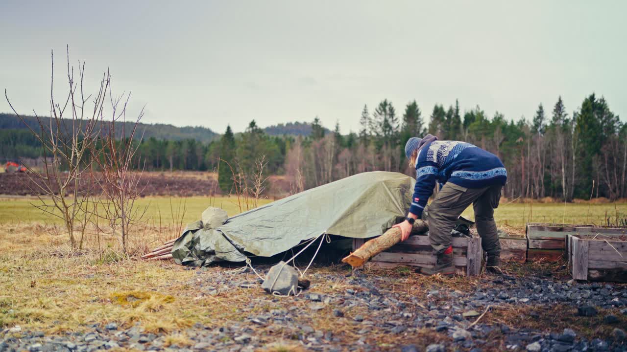 A Man Unfolds a Tarp Covering Stacks of Charred Poles - Static Shot