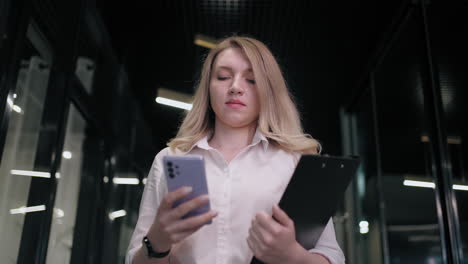 mujer de negocios sonriente mirando el teléfono celular en el interior. mujer de negocios sorprendida leyendo un mensaje en el teléfono móvil en el pasillo de la oficina. mujer mirando la pantalla del teléfono inteligente en el centro de negocios. oficina caminando