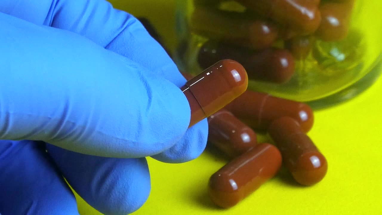 Doctor's gloved hand holding a brown medication capsule from a glass bottle on a yellow background