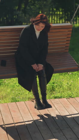 una dama con un elegante abrigo negro y botas se sienta en un columpio de madera en un parque iluminado por el sol, mirando serenamente a la distancia, el teléfono en la silla, la valla de hierro en el fondo