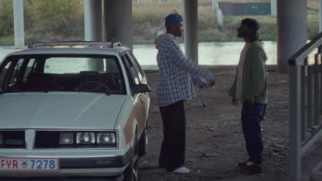 two black men fervently talking by car under overpass in gritty urban area