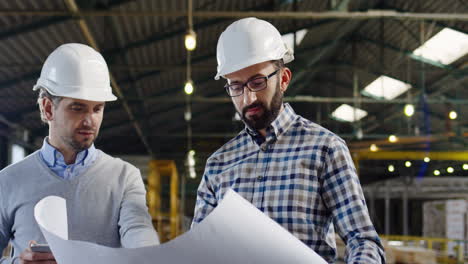 two caucasian men wearing helmets talking and looking at a blueprint in a factory