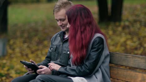 Cute-couple-sitting-on-a-bench-in-autumn-park-and-using-a-digital-tablet-together.-Young-woman-with-red-hair-and-attractive-man-in-a-leather-jacket-discussing-someting