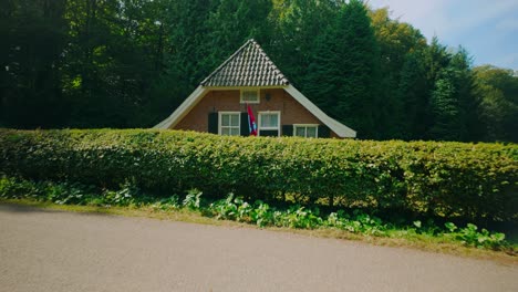 a traditional dutch farmhouse with a pointed roof sits behind a neatly trimmed hedge, surrounded by lush greenery in a serene agricultural landscape. location: netherlands