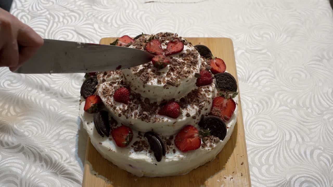 성인이 된 여성은 집 주방의 나무 절단판에 큰 주방 칼로 딸기 조각으로 아름다운 크림 케이크를 잘라 집 주방에서 휴일의 개념. 근접. 얼굴없는.  뷰.