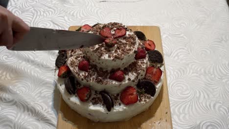 una mujer adulta corta un hermoso pastel de crema con rebanadas de fresa con un gran cuchillo de cocina en una tabla de cortar de madera en una cocina casera. el concepto de las vacaciones. primer plano. sin rostro. vista superior. 4k.