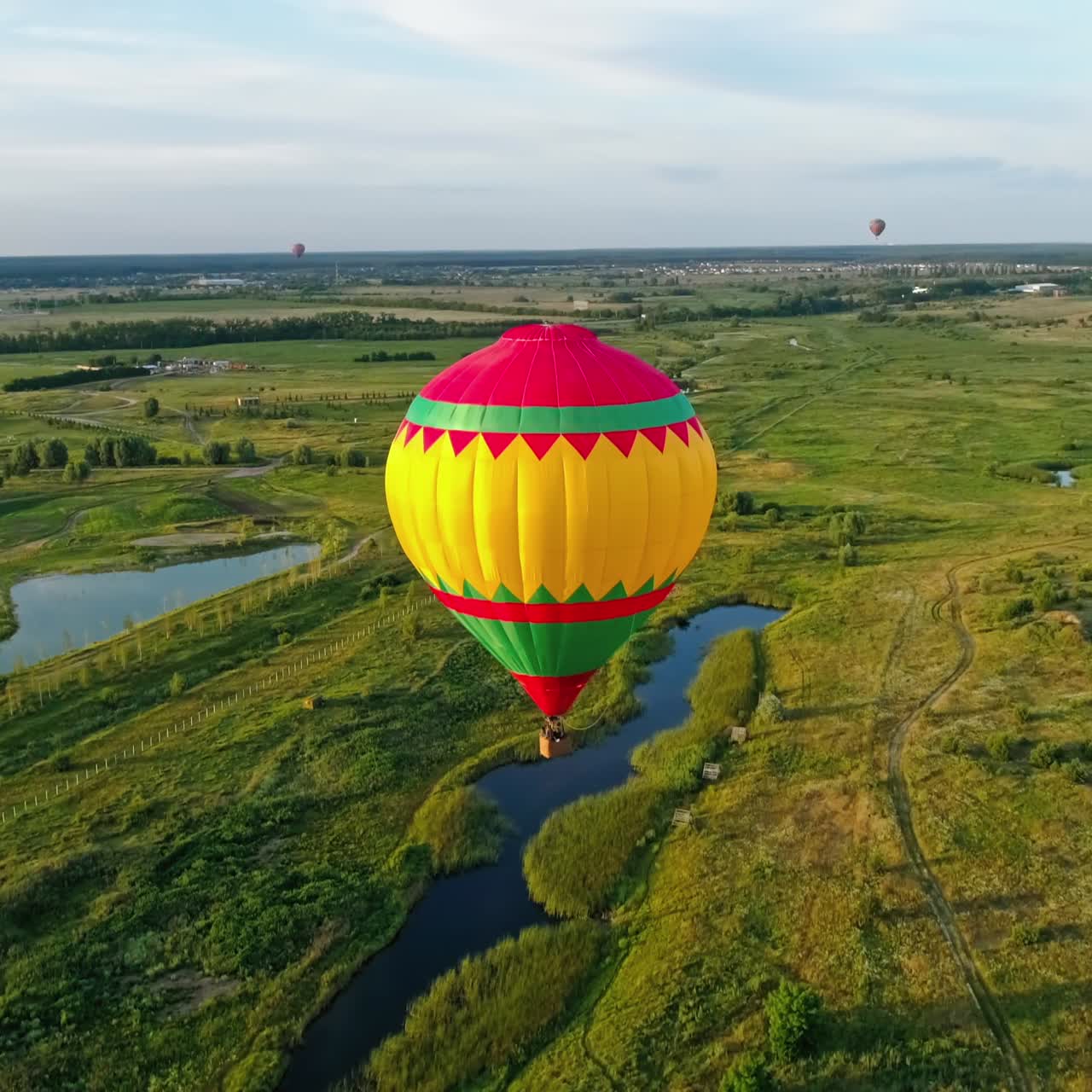 Colorful aerostat. Beautiful hot air balloon travels in the air on the background of green fields and small lakes in the rural place. Aerial view.