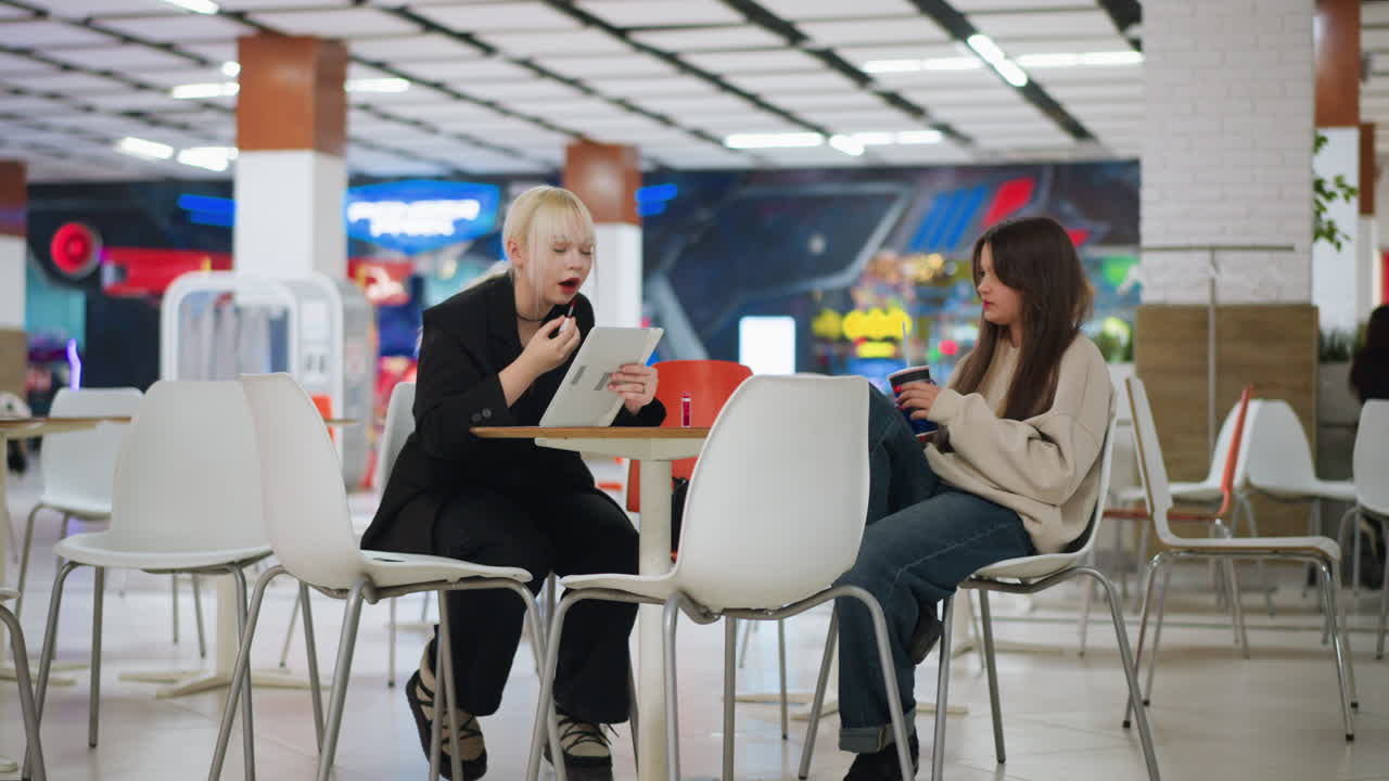 Two young women sitting in modern cafe as one applies lipstick using tablet mirror while other holds drink sipping, showing casual sisterly bond, focus on beauty routine